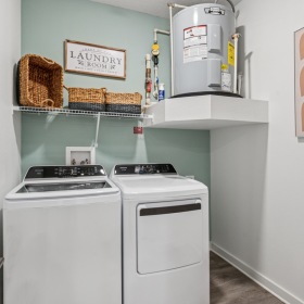 Laundry room inside a Momentum Blanding apartment featuring a washer, dryer, overhead storage shelves with baskets, and a mounted water heater.